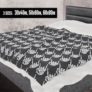 May include: A black and white throw blanket with a repeating pattern of skeleton hands making the rock and roll hand gesture. The blanket is shown on a bed with white sheets and a black headboard. The text "3 SIZES: 30x40in, 50x60in, 60x80in" is displayed at the top of the image.