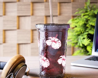 May include: A clear plastic tumbler with a straw and a dark liquid inside. The tumbler has a white hibiscus flower design. The tumbler is sitting on a wooden surface.
