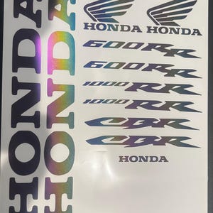 May include: A sheet of adhesive decals featuring the Honda logo, the text Honda, 600RR, 1000RR, and CBR in a holographic, iridescent finish that shifts between blue, purple, green, and yellow tones against a plain white background.