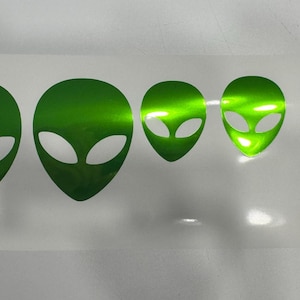 Pode incluir: Uma folha de autocolantes de cabeças de alienígenas verdes em vários tamanhos. Os autocolantes têm um acabamento metálico e estão dispostos em fila, com o maior à esquerda e diminuindo de tamanho para a direita. As cabeças dos alienígenas têm olhos grandes e ovais.