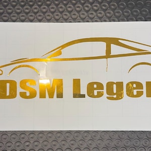 Może przedstawiać: Złota naklejka z sylwetką samochodu i napisem "DSM Legend" z logo Instagram. Projekt znajduje się na białym prostokątnym tle.