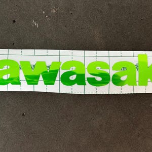 Puede incluir: Letras de vinilo verde deletreando la palabra "Kawasaki" sobre un fondo blanco cuadriculado.
