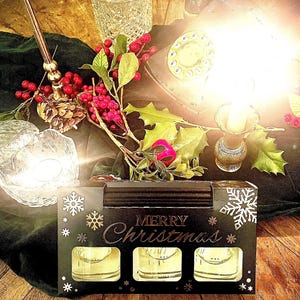 May include: A black box with the words "Merry Christmas" on it. The box contains three clear glass candle holders with gold accents. The box is sitting on a wooden table with a green plant, a glass decanter, and a vintage rotary phone.