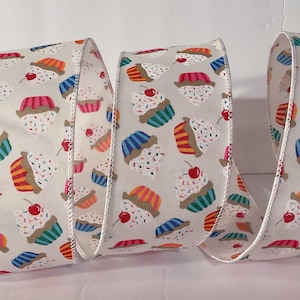 May include: A wide decorative ribbon featuring a repeating pattern of cupcakes with white frosting, colourful sprinkles, and a red cherry on top. The cupcakes have various coloured liners including blue, orange, and pink, set against a light beige background.