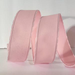 May include: A continuous length of light pink ribbon featuring a smooth, solid-coloured surface and delicate, scalloped white edges along both sides. The ribbon is arranged in a series of soft, flowing loops, showcasing its consistent texture.