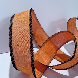 May include: A length of orange ribbon with a textured, woven appearance and a distinct black stitched edge. The ribbon is coiled in a loose, flowing shape, showcasing the contrast between the vibrant orange fabric and the dark, defined border.