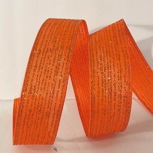 Könnte beinhalten: Ein leuchtend orangefarbenes Band mit einer strukturierten Oberfläche und vertikalen Streifen aus schimmerndem Goldglitzer. Das Band ist in einer Reihe von weichen, fließenden Schleifen angeordnet, die den orangefarbenen Stoff und den Glanz betonen.