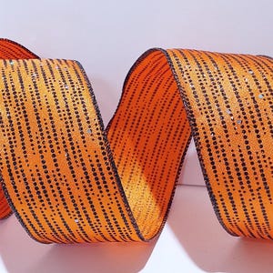 Könnte beinhalten: Ein leuchtend orangefarbenes Band mit einem sich wiederholenden Muster aus vertikalen schwarzen gepunkteten Linien und dezenten silbernen Glitzerakzenten. Das Band ist in fließenden, wellenförmigen Kurven angeordnet, die seine Textur betonen.