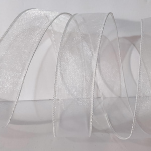 May include: A translucent white ribbon with a shimmering, glittery texture throughout the centre. The ribbon features a solid, opaque white finished edge on both sides and is arranged in a series of flowing, vertical loops and curves.