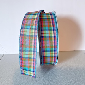 May include: A spool of colourful plaid ribbon. The ribbon features a pattern of intersecting lines in shades of pink, blue, green, orange, and yellow. The edges are trimmed with a teal blue border.