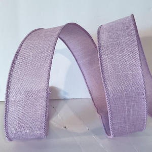 May include: A lavender-coloured ribbon with a woven, textured fabric appearance. The ribbon features a distinct, slightly darker stitched edge along both sides and is arranged in a loose, flowing S-curve shape against a plain white background.