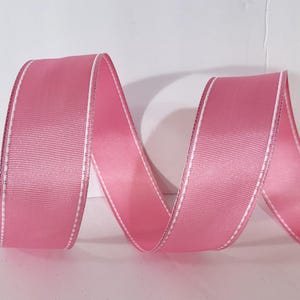 May include: A solid pink ribbon with a smooth, ribbed texture and a thin, white stitched border running along both edges. The ribbon is arranged in a series of soft, flowing curves, creating a continuous wave-like pattern across the frame.