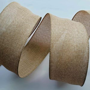 Pode incluir: Um rolo de fita de juta larga, bege. A fita é bem tecida com uma textura visível e uma borda ligeiramente mais escura. A fita é enrolada e torcida, mostrando sua largura e textura. Ideal para artesanato e decoração.