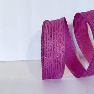 May include: A vibrant magenta ribbon featuring a textured surface with a subtle glittery finish. The ribbon is coiled into a loose spiral shape, showcasing its deep pink hue and the fine, shimmering particles embedded across the fabric material.