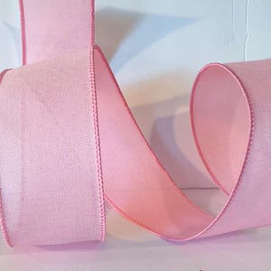 May include: A wide, light pink ribbon with a woven, textured fabric appearance and finished edges. The ribbon is curled into a loose, flowing shape, displaying its consistent pastel pink color throughout the entire length of the material.