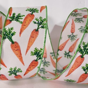 May include: A white wired ribbon featuring a repeating pattern of orange carrots with green leafy tops. The ribbon has a finished green stitched edge and is arranged in a series of soft, flowing folds that display the carrot print throughout.