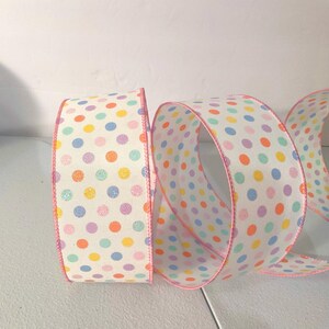 Puede incluir: Rollo de cinta decorativa con fondo blanco y lunares coloridos en tonos amarillo, naranja, rosa, morado y azul. Los bordes de la cinta están ribeteados con un fino borde rosa. Ideal para manualidades y envolver regalos.