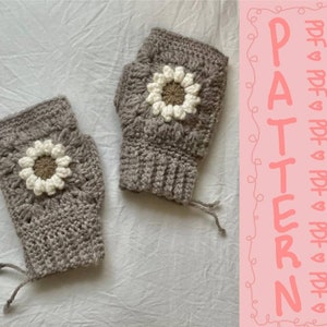 May include: A pair of grey crocheted fingerless gloves with white crocheted flowers. The gloves are on a white surface. The text "PATTERN" is written in pink on a pink background.