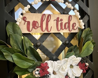 Op de afbeelding: Een ronde krans met een houten bordje met de tekst roll tide in rode letters. Het frame is versierd met witte en zwarte houten kralen, groene magnoliabladeren, witte magnoliabloemen, kleine rode bloemaccenten en een zwart-wit geblokte bal.