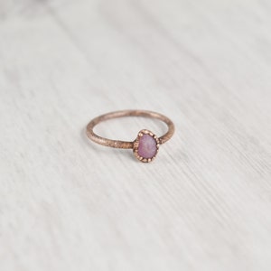 May include: A delicate ring with a textured band and a small, pink gemstone set in a bezel setting.