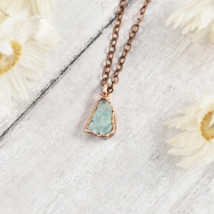 May include: A necklace featuring a raw, light blue-green stone pendant encased in a copper-toned metal setting, suspended from a matching copper-toned metal chain with oval links. The pendant has an irregular, angular shape with a textured finish.