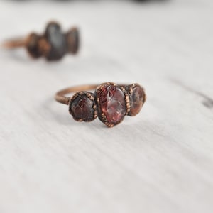 May include: A copper ring with three red gemstones set in the band. The ring is on a white surface.