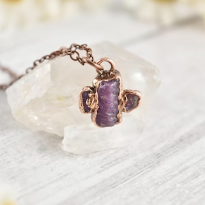May include: A necklace featuring a raw, deep purple gemstone pendant encased in a copper-toned metal setting. The pendant is shaped like a cactus with a central vertical piece and two smaller side protrusions. It hangs from a thin copper-toned chain.