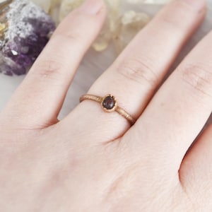 May include: A simple gold ring with a small, dark red gemstone set in the center. The ring is worn on a person's finger.