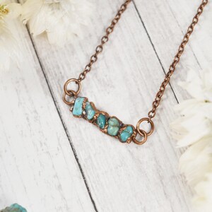 May include: A copper-colored necklace featuring a row of turquoise stones. The stones are set in a horizontal bar, connected to a delicate chain. The necklace is displayed on a white wooden surface, with white flowers in the background.