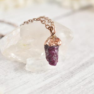 May include: A pendant necklace featuring a raw, deep purple-red gemstone shaped like an acorn, capped with a textured copper-toned metal setting. The pendant hangs from a matching copper-toned metal chain, showcasing a natural, unpolished stone texture.