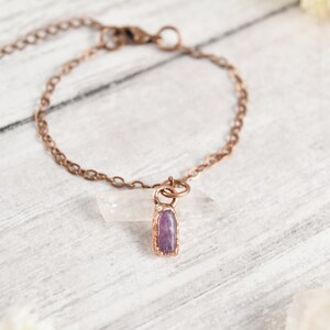 May include: A copper-toned chain bracelet featuring a single, small, rectangular purple gemstone pendant encased in a copper-colored metal setting. The bracelet includes a lobster claw clasp and an adjustable chain link extension for sizing.
