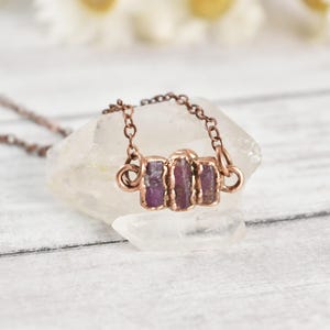 May include: A necklace featuring three small, rectangular, deep pink raw gemstone pieces set in a copper-toned metal frame. The pendant is attached to a thin, matching copper-toned metal chain with circular links, showcasing a metallic finish.