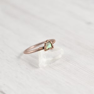 May include: A copper-colored ring with a triangular, iridescent gemstone. The ring's band has a textured, twig-like appearance, and the gemstone is set in a decorative bezel. The ring is displayed on a clear, crystalline surface.
