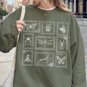 May include: Olive green sweatshirt featuring a collection of insect illustrations in a postage stamp design. The white graphic includes butterflies, bees, and other insects. The sweatshirt is worn with a baseball cap and sunglasses.