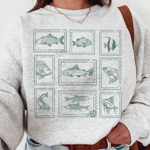 May include: A light gray sweatshirt featuring a collection of nine fish illustrations within postage stamp-style frames. The fish are depicted in a muted green color, creating a vintage aesthetic. The sweatshirt has long sleeves and a crew neck.