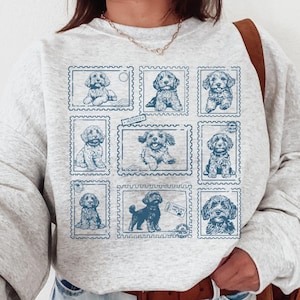 May include: A light gray sweatshirt featuring a blue postage stamp design with illustrations of various dogs. The stamps are arranged in a grid pattern across the front of the sweatshirt. The sweatshirt has long sleeves and a crew neck.