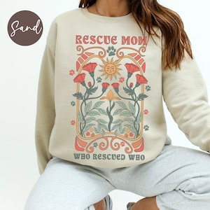 Puede incluir: Una sudadera beige con el texto "Rescue Mom Who Rescued Who" en una fuente retro. El dise&ntilde;o presenta un sol con una cara sonriente, flores y huellas de patas.