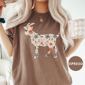 May include: Espresso-colored t-shirt featuring a floral goat design. The goat silhouette is filled with a pattern of flowers in shades of pink, blue, and cream. The shirt has a relaxed fit and short sleeves.