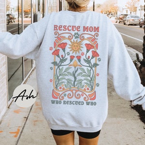 Puede incluir: Sudadera gris con un dise&ntilde;o floral y el texto "Rescue Mom Who Rescued Who" impreso en la espalda.