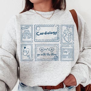 May include: A light gray sweatshirt with a blue stamp design featuring cardiology-related illustrations and the word "Cardiology." The design includes a heart, stethoscope, and EKG machine. The phrase "go with the flow" is also included.
