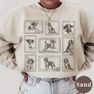 May include: Sand-colored sweatshirt featuring a grid of nine postage stamp-style illustrations of dogs in various poses. The sweatshirt is a neutral tone, and the dog illustrations are in a sepia tone. The word "Sand" is printed on a brown circle.