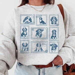 May include: A light gray sweatshirt featuring a grid of nine blue dog illustrations designed to look like postage stamps. The dogs are in various poses, and the sweatshirt has long sleeves.