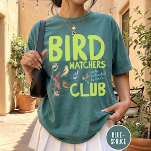 May include: A dark teal t-shirt with the text BIRD WATCHERS CLUB and the phrase easily distracted by birds printed in neon yellow. The shirt features colorful illustrations of various small birds scattered around the text.