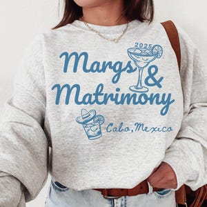 May include: A gray sweatshirt with the text "Margs & Matrimony 2025 Cabo, Mexico" in blue lettering. The design also includes a blue illustration of a margarita glass and a sombrero.
