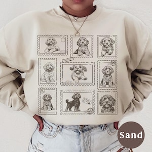 May include: Sand-colored sweatshirt featuring a grid of nine postage stamp designs, each showcasing a different sketch of a puppy. The puppies are in various poses, with detailed linework. The word "Sand" is printed on a brown circle.