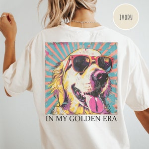 May include: A white t-shirt with a graphic of a golden retriever wearing sunglasses. The graphic is in a retro style with a colorful background. The text "IN MY GOLDEN ERA" is printed below the graphic.