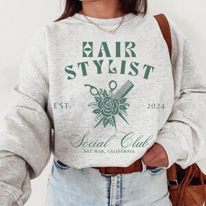 K&ouml;nnte beinhalten: Grauer Pullover mit dem Text "HAIR STYLIST" in Gr&uuml;n, einem floralen Design mit Schere und Kamm und dem Text "Social Club Del Mar, Kalifornien" in Kursivschrift.