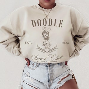 May include: A beige sweatshirt with the text "Doodle Bailey Est. 2024 Social Club Del Mar, California" and a doodle of a dog.