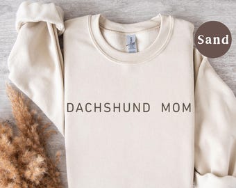 May include: A sand-colored sweatshirt with the text "DACHSHUND MOM" in black. The long-sleeved top has a crew neck and rolled cuffs. A sprig of dried ornamental grass is in the lower left corner.