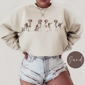 May include: A beige sweatshirt with a graphic print of five brown and white German Shorthaired Pointers. The model is wearing the sweatshirt with a pair of light blue denim shorts.